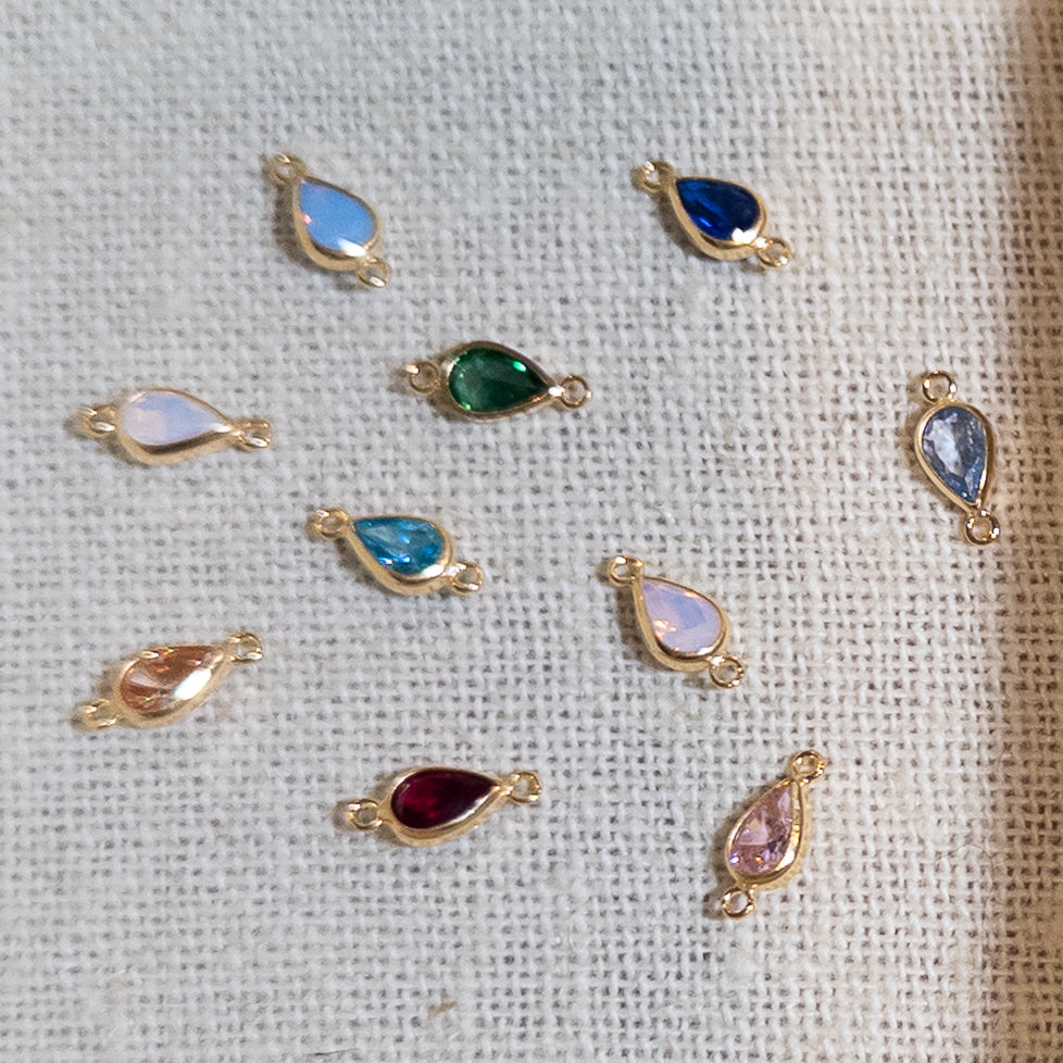 Nine gold teardrop-shaped jewelry charms with different colored stones are arranged on a textured beige linen fabric background.