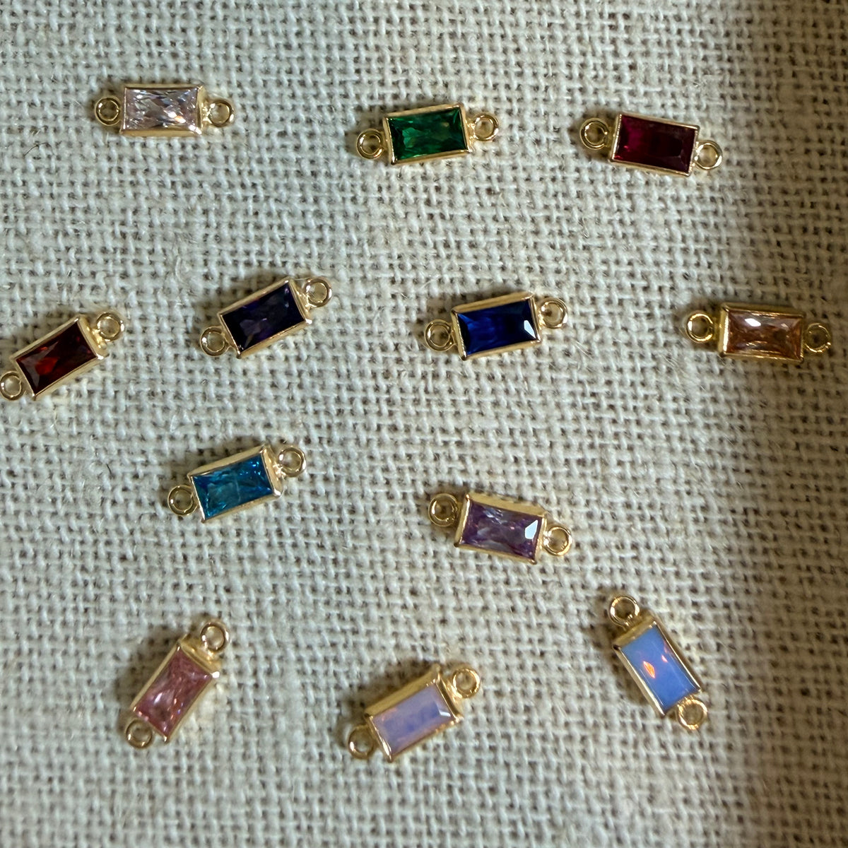 Nine small gold-tone rectangular bezel settings with colorful faceted stones, arranged on a textured off-white fabric background.