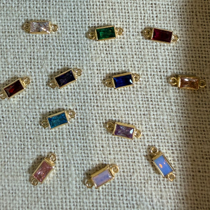 Nine small gold-tone rectangular bezel settings with colorful faceted stones, arranged on a textured off-white fabric background.